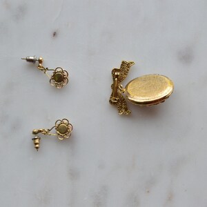 Puede incluir: Juego de joyas doradas con dos pendientes de bot&oacute;n en forma de flor y un peque&ntilde;o relicario ovalado. Los pendientes tienen un dise&ntilde;o delicado con una piedra verde central. El relicario tiene una superficie texturizada.