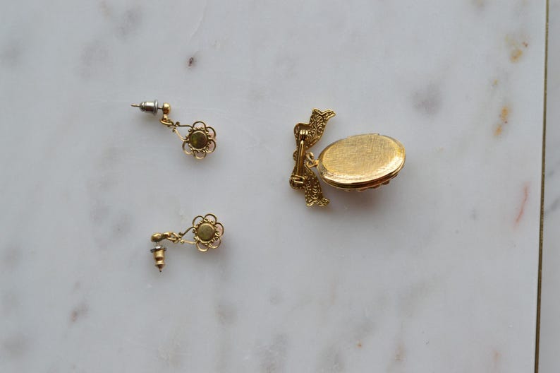 Puede incluir: Juego de joyas doradas con dos pendientes de bot&oacute;n en forma de flor y un peque&ntilde;o relicario ovalado. Los pendientes tienen un dise&ntilde;o delicado con una piedra verde central. El relicario tiene una superficie texturizada.