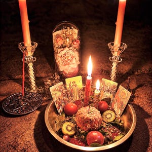 May include: A mystical arrangement featuring lit candles, tarot cards, and a bowl of fruit. Two tall candles flank a central display with a glass dome containing flowers. The scene is illuminated with warm light, creating a spiritual ambiance.
