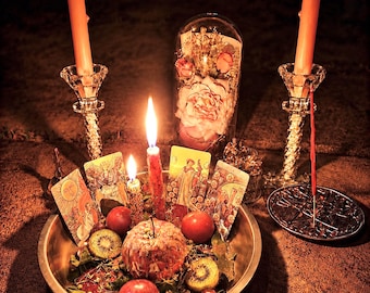 May include: A still life arrangement featuring lit candles, tarot cards, and a bowl of fruit. Two tall candles flank the scene, with a central bowl holding apples, kiwi, and other items. A glass dome with flowers is in the background.