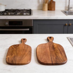 May include: Two wooden chopping boards with handles. The boards are rectangular with rounded corners and a natural wood grain finish. One board is slightly smaller than the other. Kitchenware.