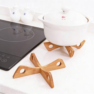May include: Two bamboo trivets with a white ceramic pot on one. The trivets are made of light brown wood and have a cross-like design with circular cutouts. The pot has a lid and sits on a trivet on a white countertop.