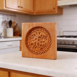 Könnte beinhalten: Eine Holzkeksform mit floralem Design. Die quadratische Form zeigt ein kreisförmiges Muster aus Rosen und Blättern, das in das Holz geschnitzt ist. Die Form ist hellbraun und steht auf einer weißen Arbeitsplatte in einer Küche.