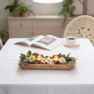 Peut inclure: Un centre de table en bois avec deux bougies allumées dans des bocaux en verre, entourées de fleurs blanches et crème, de verdure et de pierres décoratives. Le plateau est posé sur une nappe blanche.