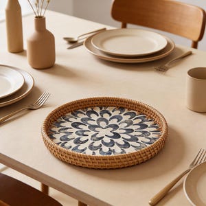 May include: A round woven tray with a blue and white floral design sits on a light-colored table. The tray is surrounded by tableware, including plates, forks, and a mug. A vase with dried stems is also visible.