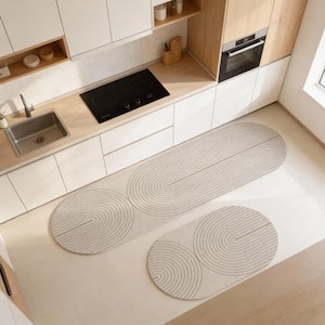 May include: Two beige rugs with a geometric design in a kitchen. The larger rug is oval, and the smaller is round. The rugs feature concentric semi-circles and straight lines. The kitchen has white cabinets and wooden accents. The rugs are likely made of a soft material.