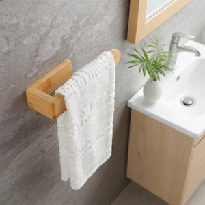 May include: A bamboo towel holder mounted on a gray wall, holding a white towel. The towel has a textured pattern. The holder is made of light-colored bamboo and has a rectangular design. A bathroom sink and mirror are in the background.