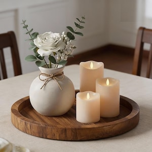 Może przedstawiać: Dekoracyjna drewniana taca z białym wazonem z białą różą i zielenią oraz trzema kremowymi świecami LED. Świece mają różne rozmiary i efekt migoczącego płomienia. Kompozycja stoi na stole.