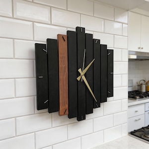 May include: A modern wall clock with a geometric design. The clock features black wooden slats arranged in a diamond shape, with a central brown slat. Gold-colored hands indicate the time. The clock is mounted on a white tiled wall.