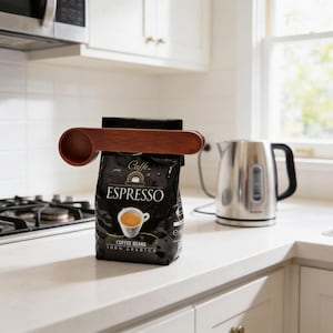 May include: A dark brown wooden coffee scoop and bag clip set. The clip is attached to a black bag of espresso coffee beans. The bag has the word "ESPRESSO" in white lettering. A stainless steel electric kettle is in the background.