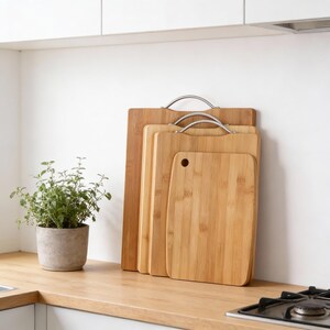 May include: A set of four bamboo cutting boards of varying sizes, with stainless steel handles. The boards are light brown and arranged vertically. A small potted plant sits to the left on a wooden countertop.