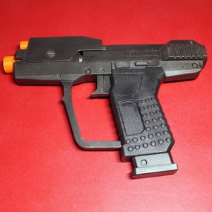 Puede incluir: Una pistola de juguete negra y gris con puntas naranjas. La pistola tiene un agarre texturizado y un diseño futurista. La imagen está sobre un fondo rojo.