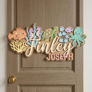 Pode incluir: Uma placa de porta de madeira com tema subaquático, com um peixe-palhaço, cavalos-marinhos, uma tartaruga e um polvo. O nome "Finley" está em itálico, com "JOSEPH" abaixo, tudo em um design em camadas.