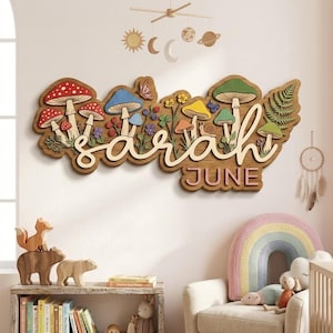 Puede incluir: Arte de pared de madera con el nombre "Sarah" en escritura cursiva y el mes "Junio" debajo. El diseño incluye setas coloridas, flores y un helecho, creando una pieza de decoración de guardería caprichosa e inspirada en la naturaleza.