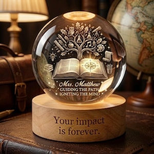Puede incluir: Un globo de cristal transparente con un diseño de árbol grabado, que incluye un libro abierto, una brújula e iconos escolares. El globo se asienta sobre una base de madera con la inscripción "Your impact is forever". El globo está iluminado desde dentro.