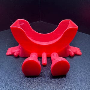 May include: A bright red, 3D-printed novelty item. The design features a curved upper section, two rounded feet, a small central protrusion, and two small hands. The item is made of plastic and sits on a dark surface.