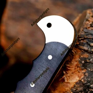 Pode incluir: Um cabo de faca com um refor&ccedil;o de metal polido cor de prata com um orif&iacute;cio circular e uma empunhadura de madeira escura texturizada fixada por dois rebites cor de prata. O texto Adisharpedges &eacute; vis&iacute;vel na superf&iacute;cie do cabo.