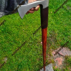 Pode incluir: Um tomahawk com uma cabeça de metal prateada, um punho envolto em couro castanho escuro e um cabo de madeira longo e cónico. O texto Adisharpedges é visível várias vezes na imagem, sobrepondo-se à cabeça de metal e ao cabo de madeira.