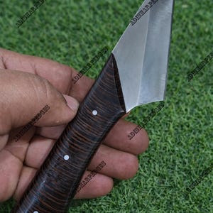 Pode incluir: Uma faca artesanal com uma lâmina de aço inoxidável e um cabo de madeira escura. O cabo apresenta três incrustações brancas. A faca é segurada contra um fundo gramado verde.