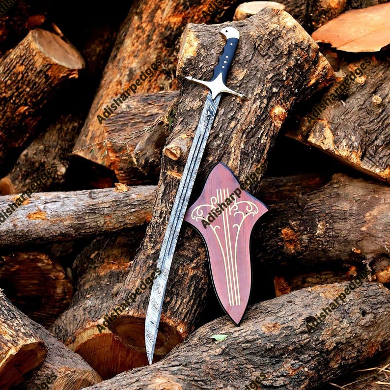 Pode incluir: Uma espada longa com l&acirc;mina de a&ccedil;o Damasco estampada, cabo preto com pomo prateado e guarda cruzada prateada. Ao lado da espada est&aacute; uma placa de parede de madeira escura com um design de linhas decorativas sim&eacute;tricas de cor clara.