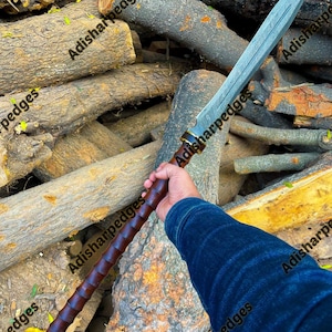 Pode incluir: Uma lança longa com uma lâmina de aço Damasco de dois gumes com um padrão ondulado visível. O cabo é feito de madeira castanha escura com uma pega segmentada e estriada, um pomo metálico de cor dourada na base e um colar dourado.