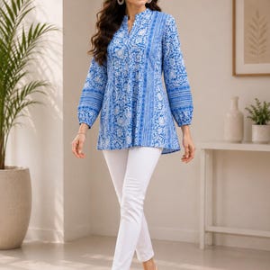 May include: A long-sleeved tunic featuring a vibrant blue and white floral pattern with a split neckline and gathered waist detail, paired with slim-fit white trousers and light-colored open-toe sandals, creating a coordinated ensemble.