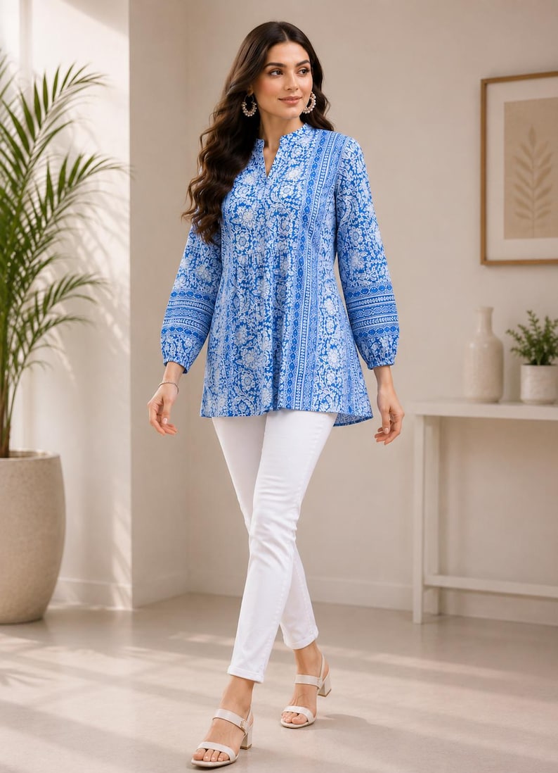 May include: A long-sleeved tunic featuring a vibrant blue and white floral pattern with a split neckline and gathered waist detail, paired with slim-fit white trousers and light-colored open-toe sandals, creating a coordinated ensemble.
