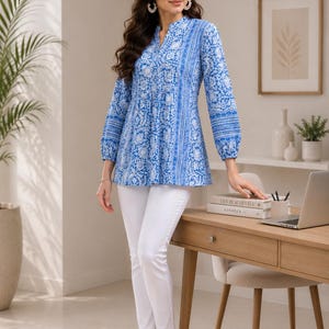 May include: A long-sleeved tunic top featuring a vibrant blue and white floral pattern with a split neckline and gathered cuffs, paired with slim-fit white trousers and light-colored open-toe sandals, creating a coordinated ensemble.