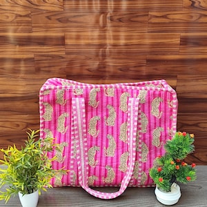 Op de afbeelding: Een roze tote bag met een herhaald luipaardprintpatroon. De tas heeft verticale strepen en witte handvatten. De achtergrond is een houten oppervlak met twee kleine potplanten aan weerszijden van de tas.
