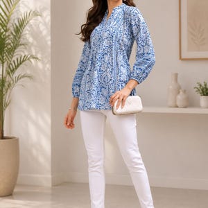 May include: A long-sleeved tunic featuring a blue and white floral block print pattern, a split V-neckline, and a gathered waistline. The tunic is paired with slim-fit white trousers, a small white quilted clutch bag, and open-toe heeled sandals.