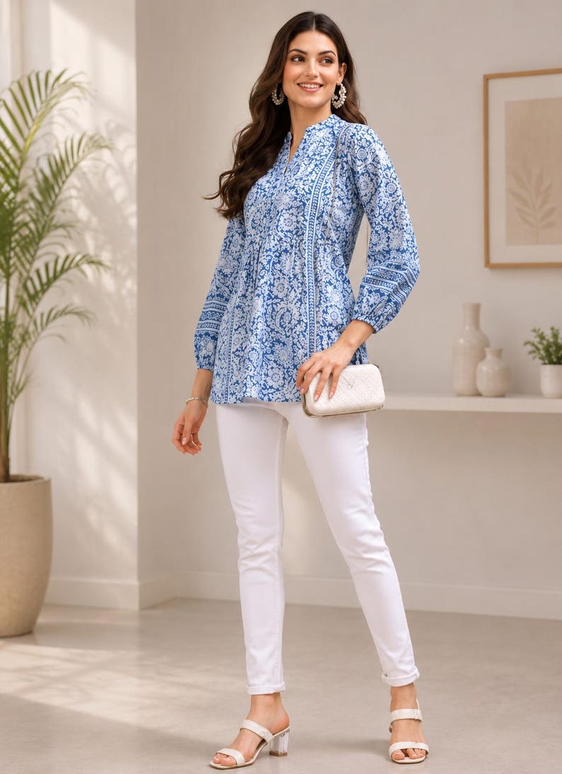 May include: A long-sleeved tunic featuring a blue and white floral block print pattern, a split V-neckline, and a gathered waistline. The tunic is paired with slim-fit white trousers, a small white quilted clutch bag, and open-toe heeled sandals.