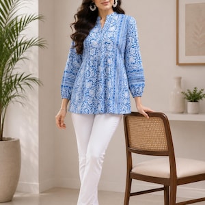 May include: A long-sleeved tunic top featuring a blue and white floral block print pattern with a split V-neckline and gathered waist, paired with solid white slim-fit trousers and white open-toe sandals with a low block heel.