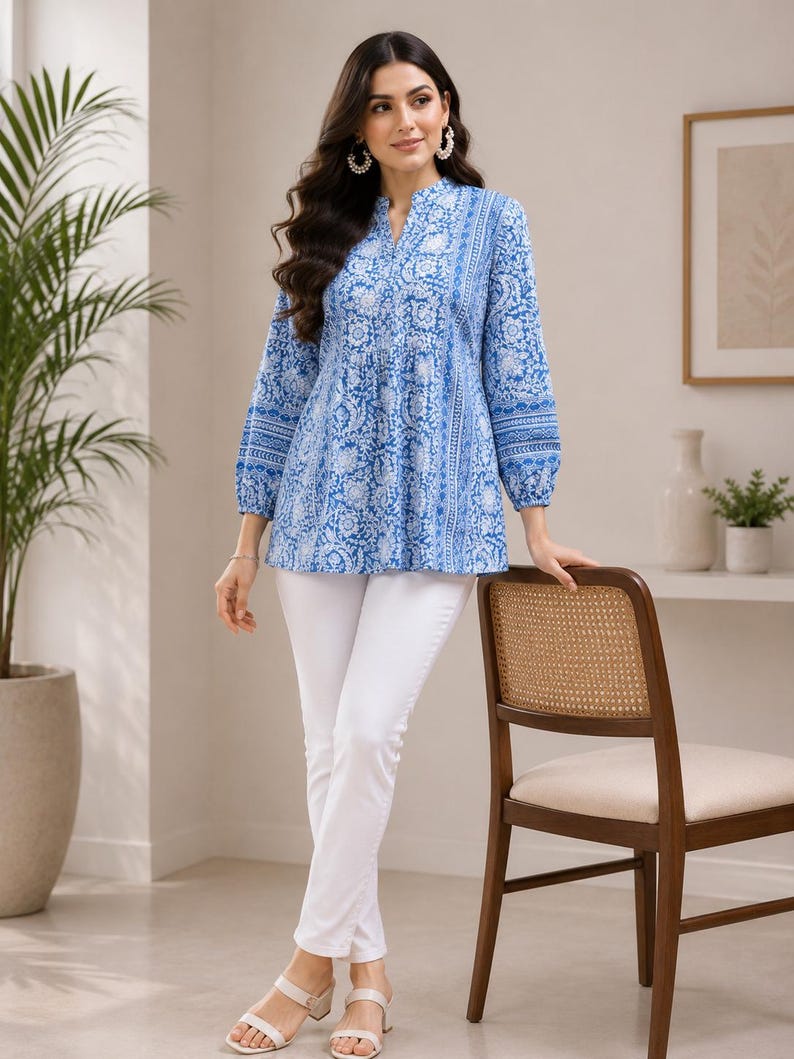 May include: A long-sleeved tunic top featuring a blue and white floral block print pattern with a split V-neckline and gathered waist, paired with solid white slim-fit trousers and white open-toe sandals with a low block heel.