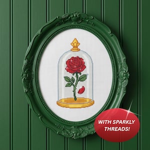 May include: A cross-stitch embroidery of a red rose inside a glass cloche with a gold base and topper, framed in an oval dark green ornate frame. The design features white sparkles and a red petal, with a red circular tag stating WITH SPARKLY THREADS!
