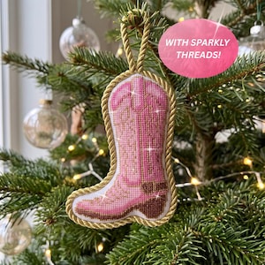 May include: A pink cowboy boot ornament featuring cross-stitch detailing and sparkly threads. The boot is outlined with a thick gold twisted rope trim and includes a matching gold rope loop at the top for hanging. Text reads: WITH SPARKLY THREADS!