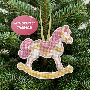 May include: A cross-stitch rocking horse ornament featuring white, pink, and gold thread with sparkly accents. The horse has a pink mane and tail, a gold saddle, and gold rockers. A gold ribbon loop is attached for hanging. Text reads: WITH SPARKLY THREADS!