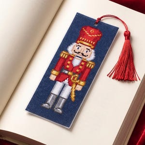 Könnte beinhalten: Ein rechteckiges Kreuzstich-Lesezeichen mit einem verpixelten Nussknacker-Soldaten-Motiv auf dunkelblauem Grund. Der Nussknacker trägt eine rote Uniform mit goldenen Akzenten und eine hohe rote Mütze. Eine rote Quaste ist oben am Lesezeichen befestigt.