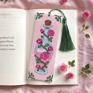 Puede incluir: Un marcapáginas rectangular de punto de cruz con una botella de cristal llena de líquido rosa, envuelta en enredaderas verdes con rosas rosas. El marcapáginas tiene una borla verde y está colocado sobre un libro abierto con texto.