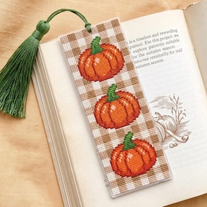 May include: A rectangular bookmark featuring a cross-stitched pattern of three orange pumpkins with green stems set against a beige and white gingham background. The bookmark is topped with a green thread tassel attached by a green cord.