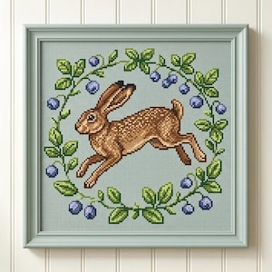 May include: A framed cross-stitch embroidery featuring a brown hare leaping in the center, surrounded by a circular wreath of green leaves and blue berries against a light blue fabric background, all encased in a light green wooden frame.