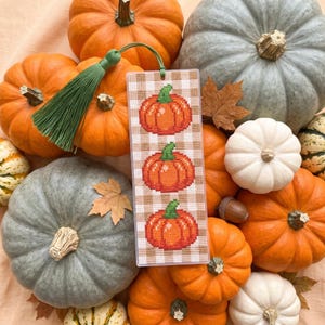 May include: A rectangular bookmark featuring a cross-stitched pattern of three orange pumpkins on a beige and white gingham background. The bookmark is encased in a clear plastic sleeve and is finished with a green tassel attached to the top.
