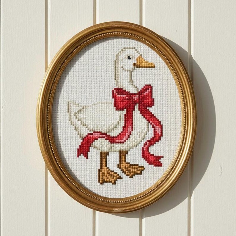 May include: Oval framed cross-stitch art featuring a white goose with a red ribbon bow. The goose has yellow feet and beak. The frame is gold-colored and the background is white.