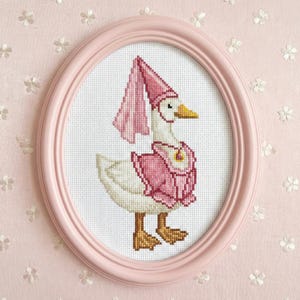 May include: A cross-stitch framed in a pink oval frame. The artwork features a white goose wearing a pink hat and vest. The goose has a yellow beak and brown feet. The background is a light pink floral pattern.