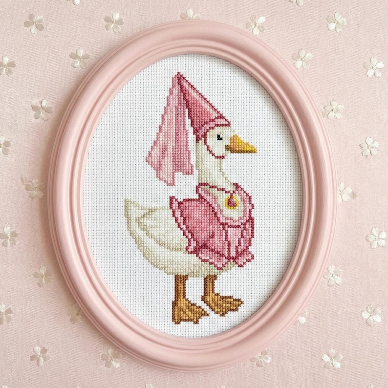 May include: A cross-stitch framed in a pink oval frame. The artwork features a white goose wearing a pink hat and vest. The goose has a yellow beak and brown feet. The background is a light pink floral pattern.