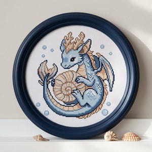 Könnte beinhalten: Ein gerahmtes Kreuzstichbild mit einem hellblauen Drachen, der sich um eine hellbraune Nautilusmuschel windet. Der Drache hat hellbraune Akzente und ist von kleinen blauen Blasen umgeben. Der runde Rahmen ist dunkelblau.