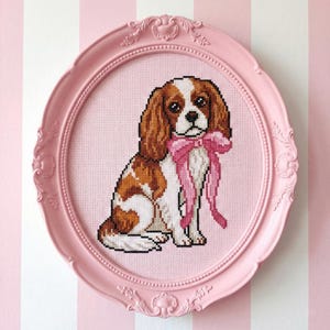 May include: A cross-stitch artwork of a Cavalier King Charles Spaniel dog wearing a pink bow, set in a pink oval frame. The dog has brown and white fur. The background is a pink and white striped pattern.