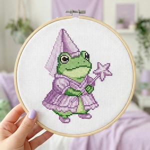 Puede incluir: Bordado de punto de cruz de una rana verde con un disfraz de mago morado y una varita en forma de estrella. El diseño está dentro de un bastidor de bordado de madera. El fondo es blanco.