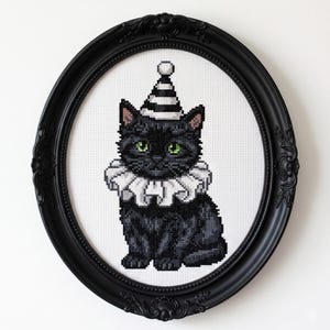 Puede incluir: Arte de punto de cruz de un gato negro con un sombrero a rayas y un cuello con volantes, enmarcado en un marco ovalado negro. El gato tiene ojos verdes y está sobre un fondo blanco.
