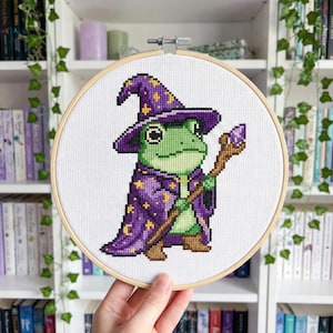 May include: Cross-stitch of a green frog wizard wearing a purple cape and hat with gold stars. The frog holds a wooden staff with a purple crystal. The cross-stitch is in a wooden embroidery hoop.