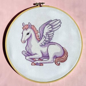 May include: A circular wooden embroidery hoop containing a cross-stitch design of a white and light purple winged horse. The horse features a pink mane and tail, with its wings spread upward, all stitched onto a white fabric background.